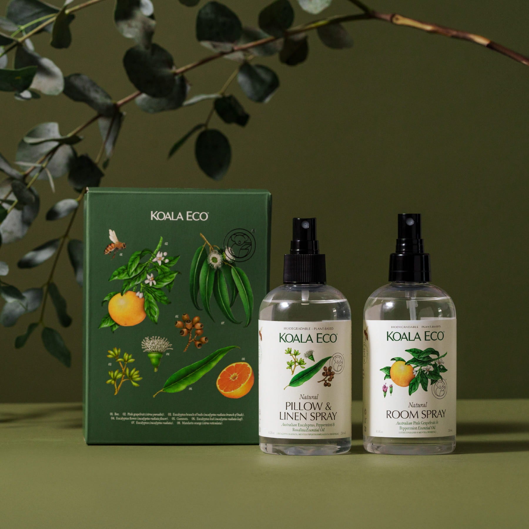 Two bottles from the "Aromatherapy Gift Collection - Room & Linen" set by Koala Eco are placed beside a coordinating ribbon-wrapped box, all against a green backdrop and tree branch over it.